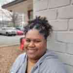 Alisa Michele, a Black woman with curly hair pulled up in back, standing outside by a painted brick wall.
